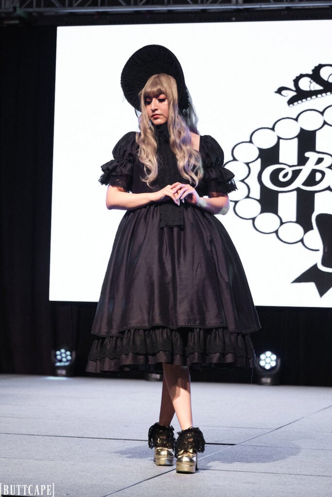 lolita model wearign all black dress and bonnet standing with legs crossed and looking down.