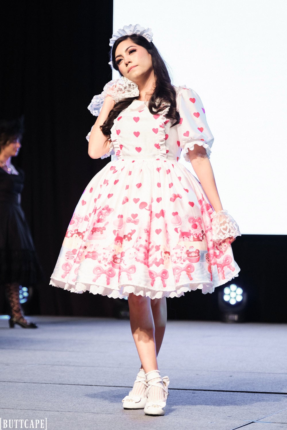 lolita model wearing white and pink dessert print dress posing with hand next to face and legs crossed.