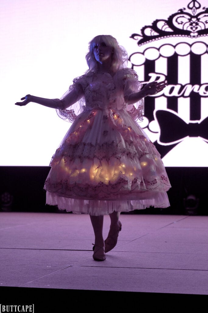 lolita model wearing lacy pink dress with LED lights in dark walking towards front of stage.