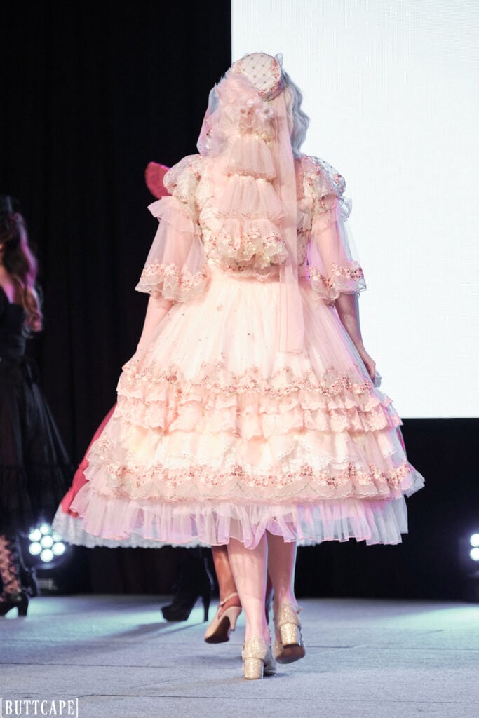 lolita model wearing lacy pink dress with LED lights walking towards back of stage.