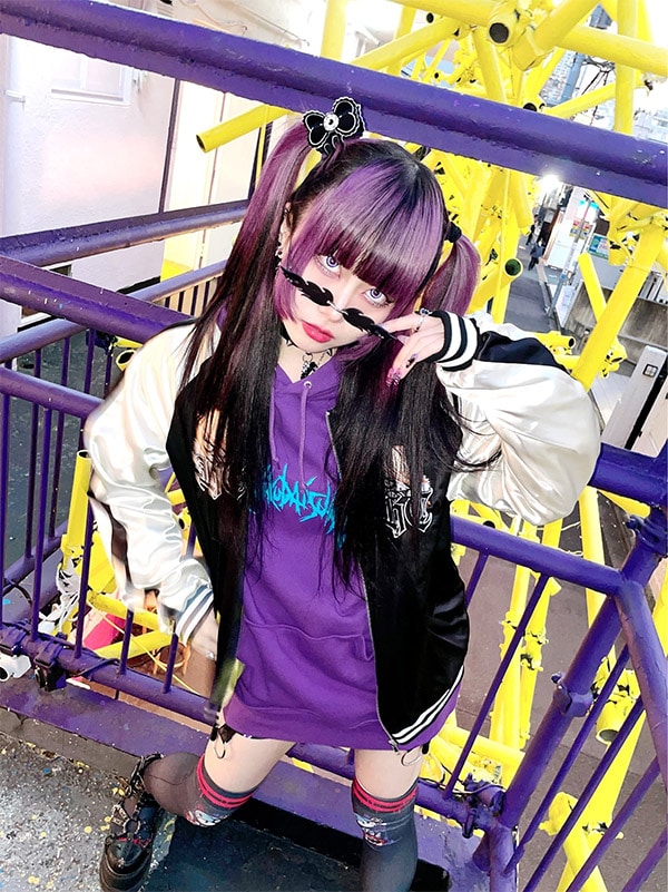 Harajuku model wearing cool sunglasses, varsity jacket, oversized hoodie, and printed over knee socks in front of colorful background.