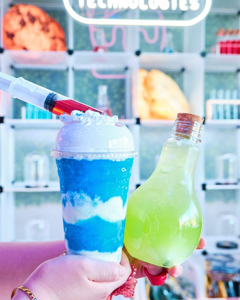 Blue raspberry and creme slushie with decorative syringe on left. Glowing green drink in lightbulb container on right.