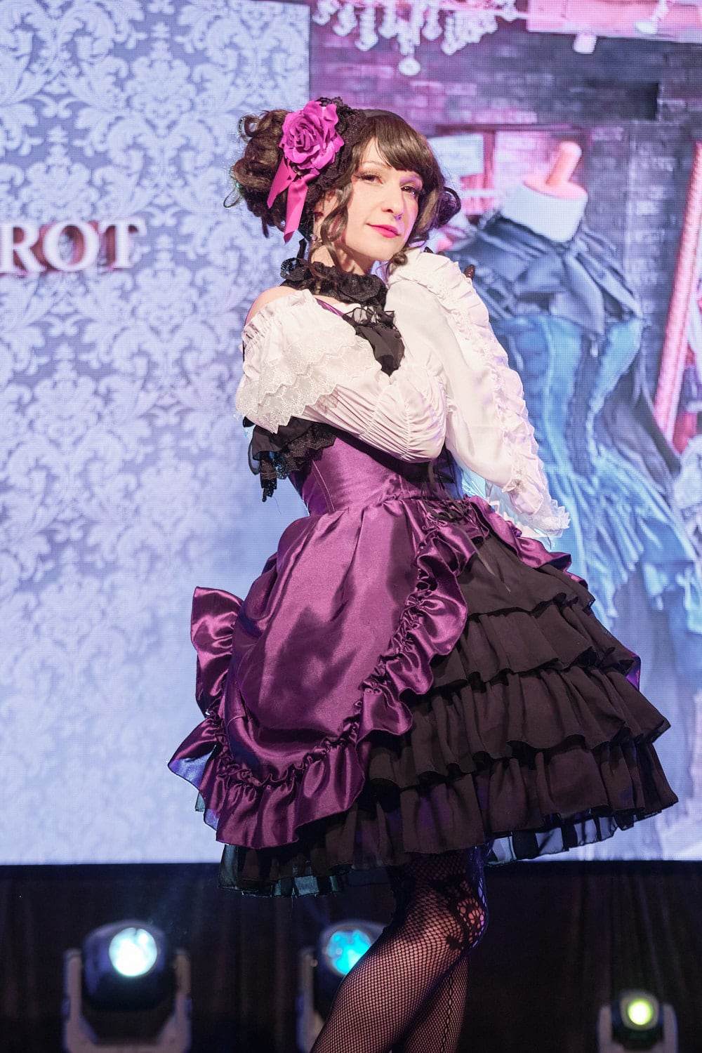 Gothic lolita wearing purple bustle dress, white princess sleeve blouse, fishnet tights; half body shot.
