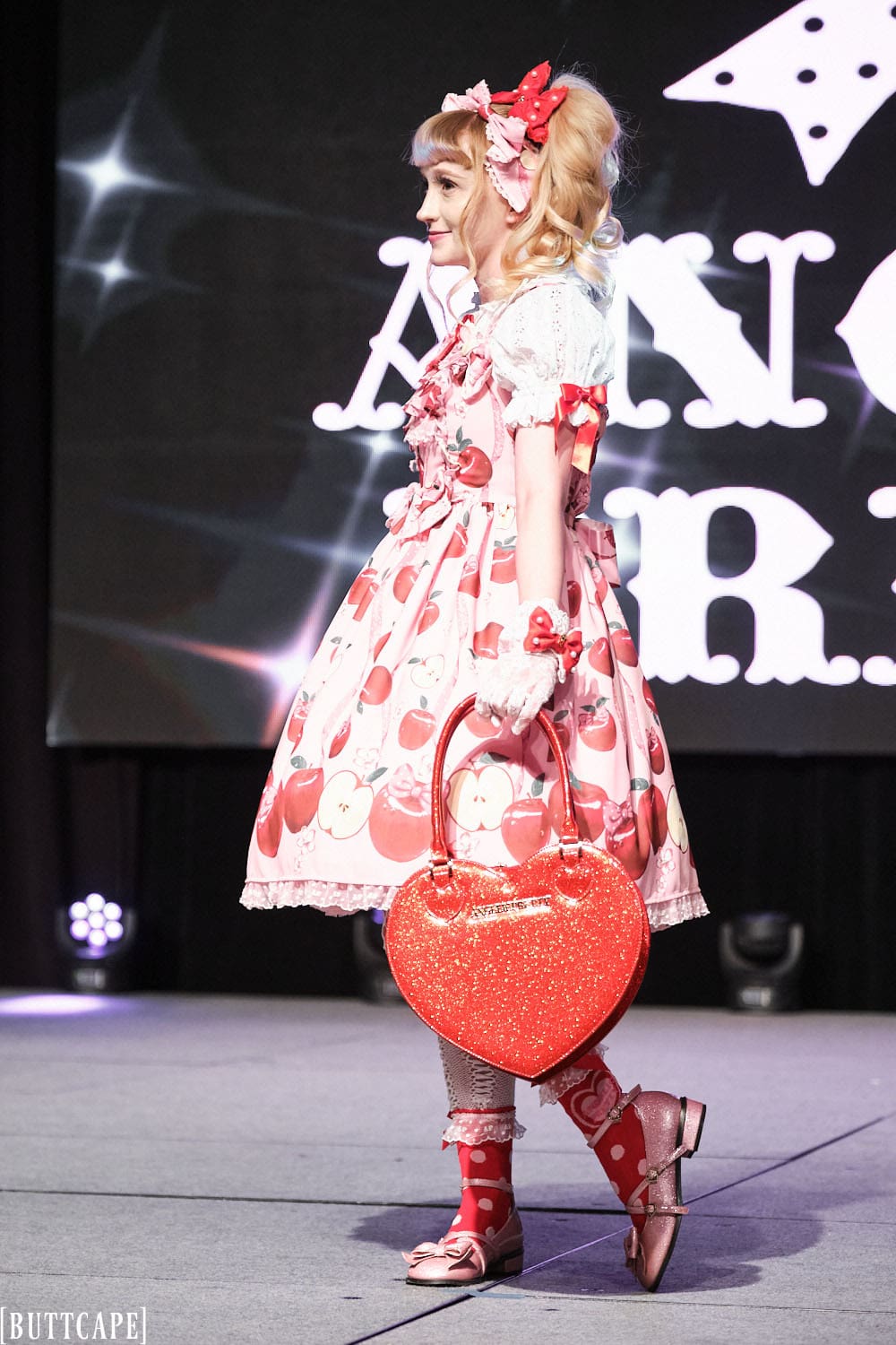 Model 3 wearing pink and red cherry themed lolita outfit holding red heart purse - side view.