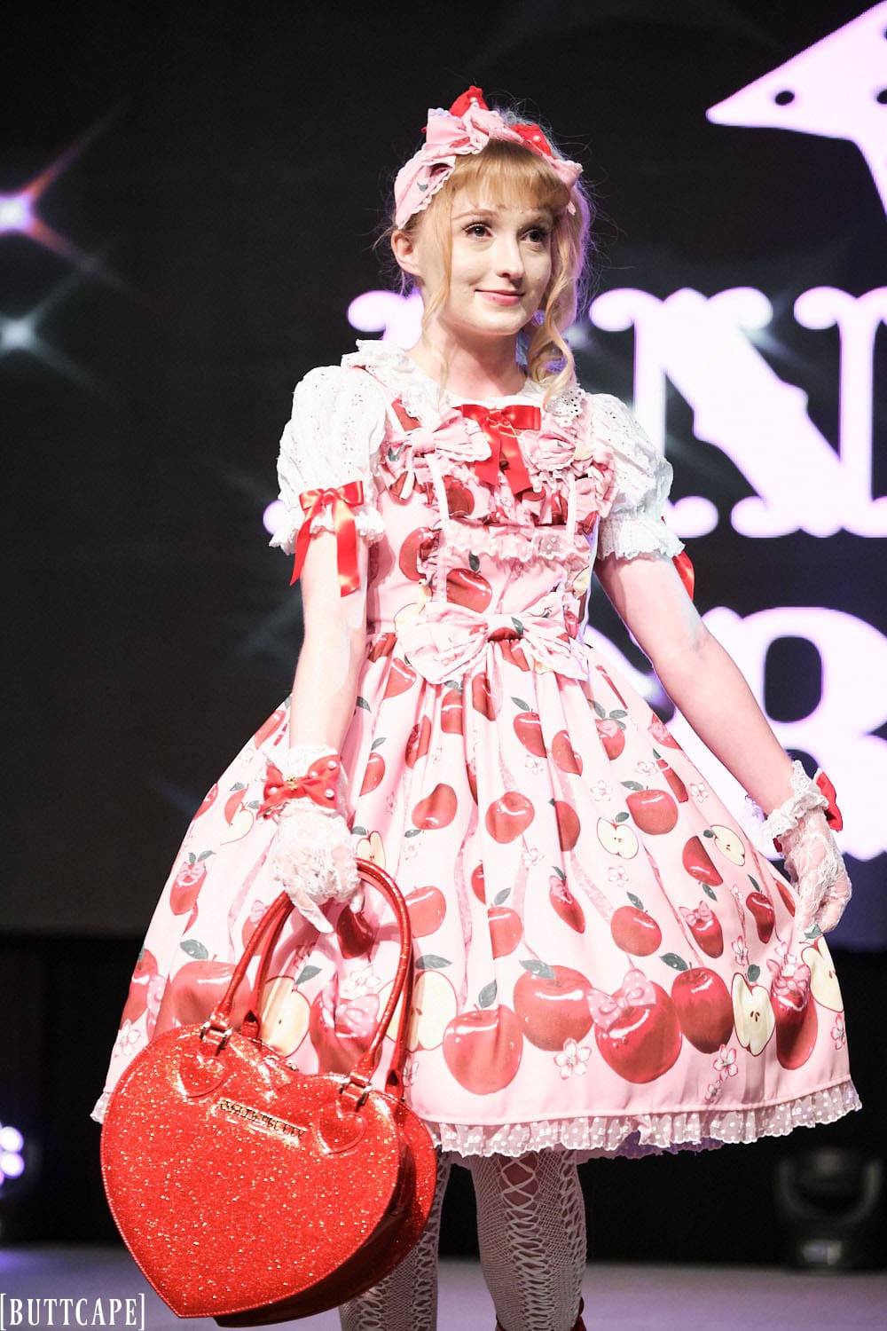 Model 3 wearing pink and red cherry themed lolita outfit holding red heart purse - half body.