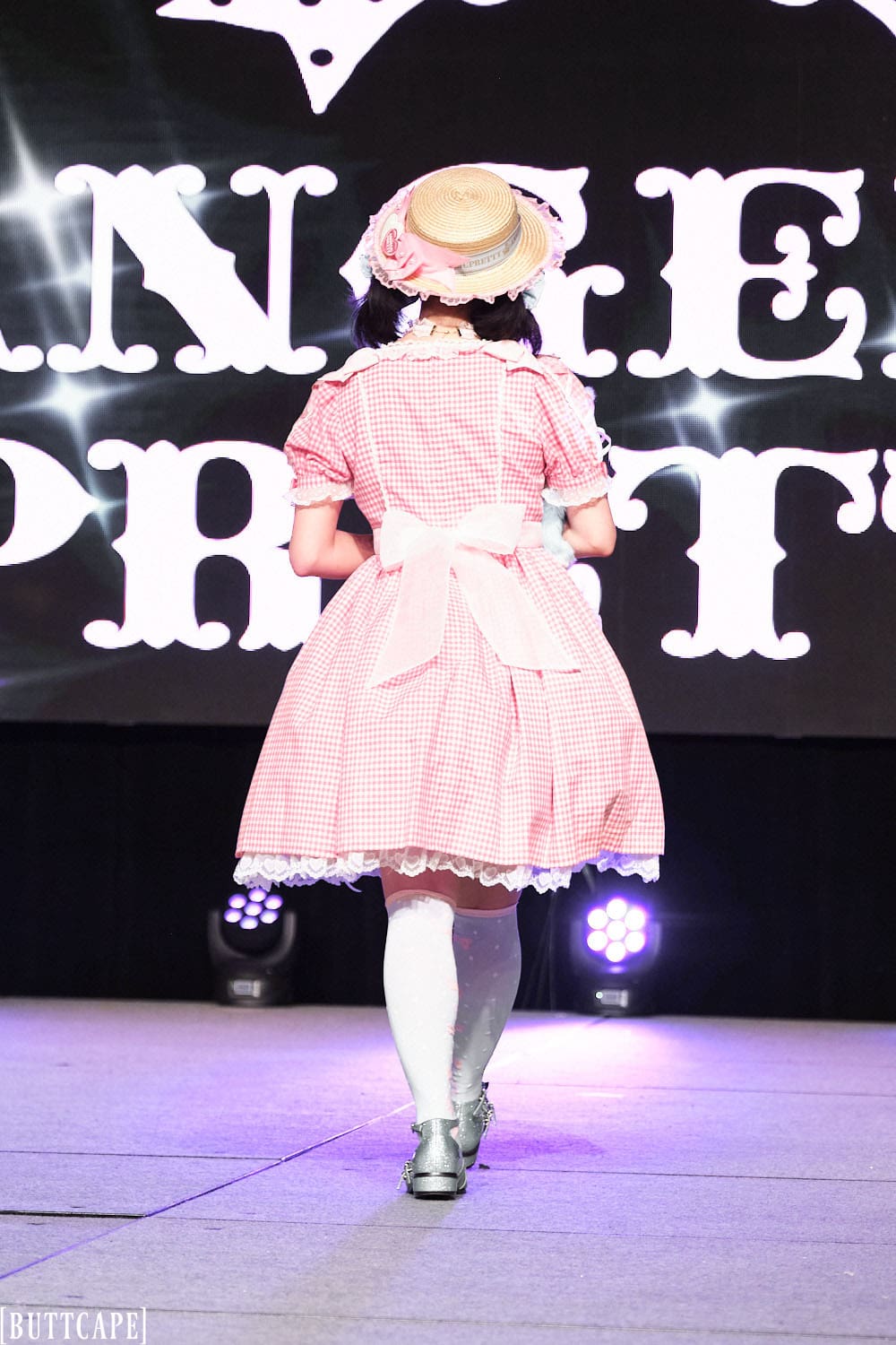 Model 11 wearing pink and white gingham lolita dress and boater hat holding two stuffed animals - back side.