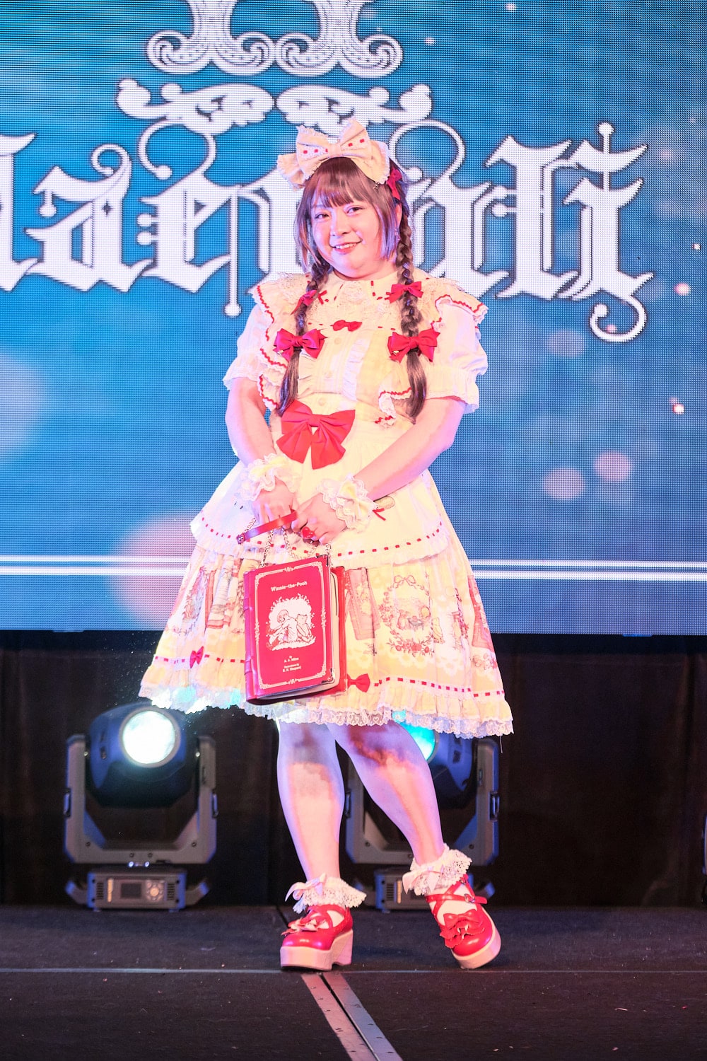 Model 3 wearing yellow and white gingham Winnie the Pooh print dress with matching headbow, half apron, and red accents holding book-shaped purse - full body 2.