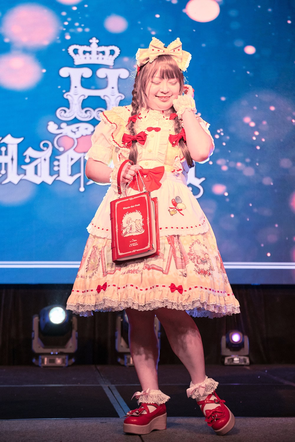 Model 3 wearing yellow and white gingham Winnie the Pooh print dress with matching headbow, half apron, and red accents holding book-shaped purse - full body 3.