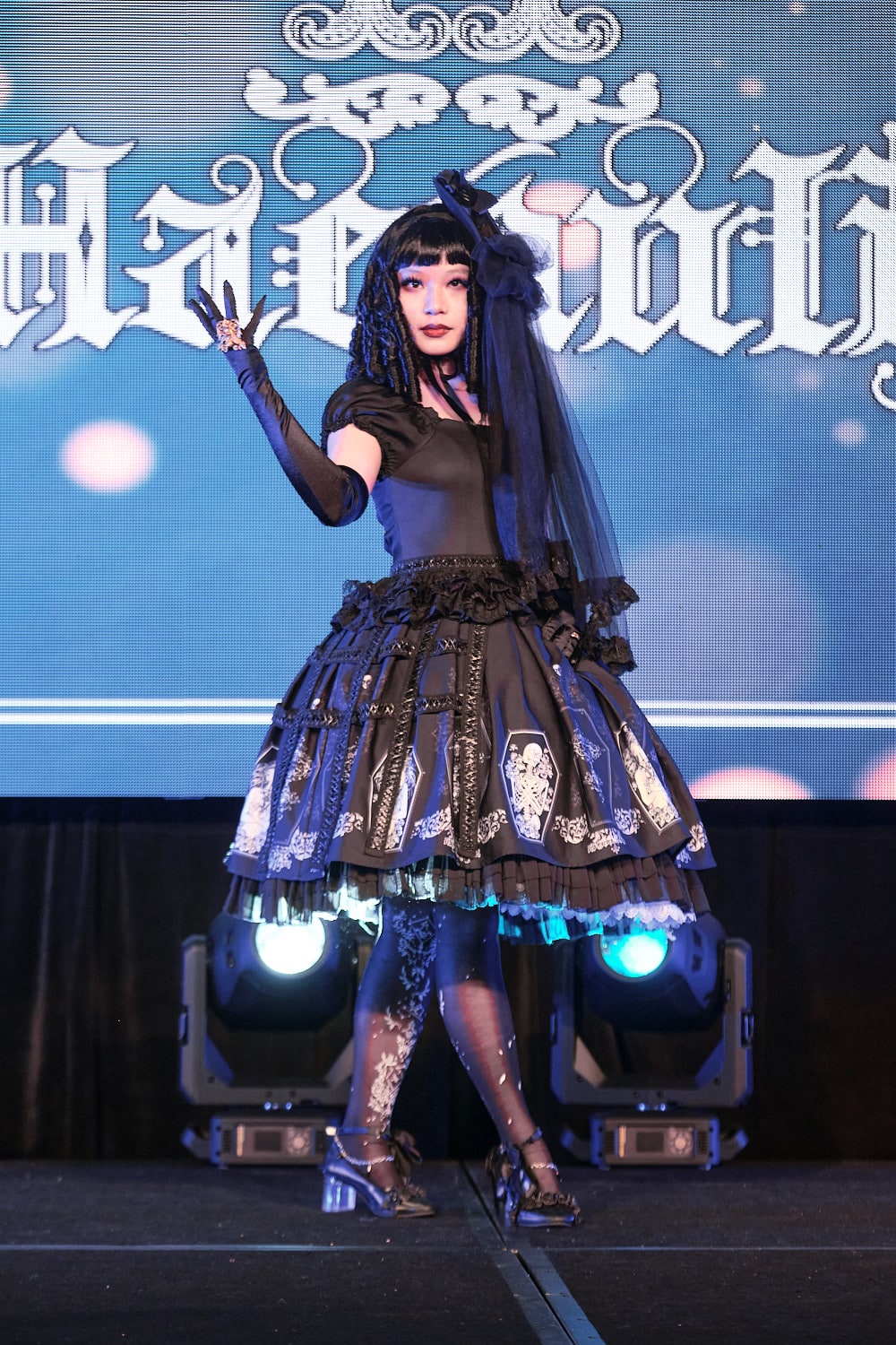 Model 10 wearing all black kuro lolita one piece dress with coffin motif border printm half cage skirt, and veil headdress - full body 1.