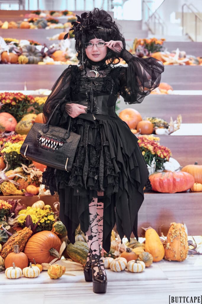 All black gothic lolita outfit featuring many layers and different fabrics. The outfit consists of a mesh blouse with dramatic sleeves, cage jumperskirt with dragon leather accents, sheer rose motif over the knee socks, platform mary jane shoes, and leather handbag with teeth motifs. Full body pose 1.
