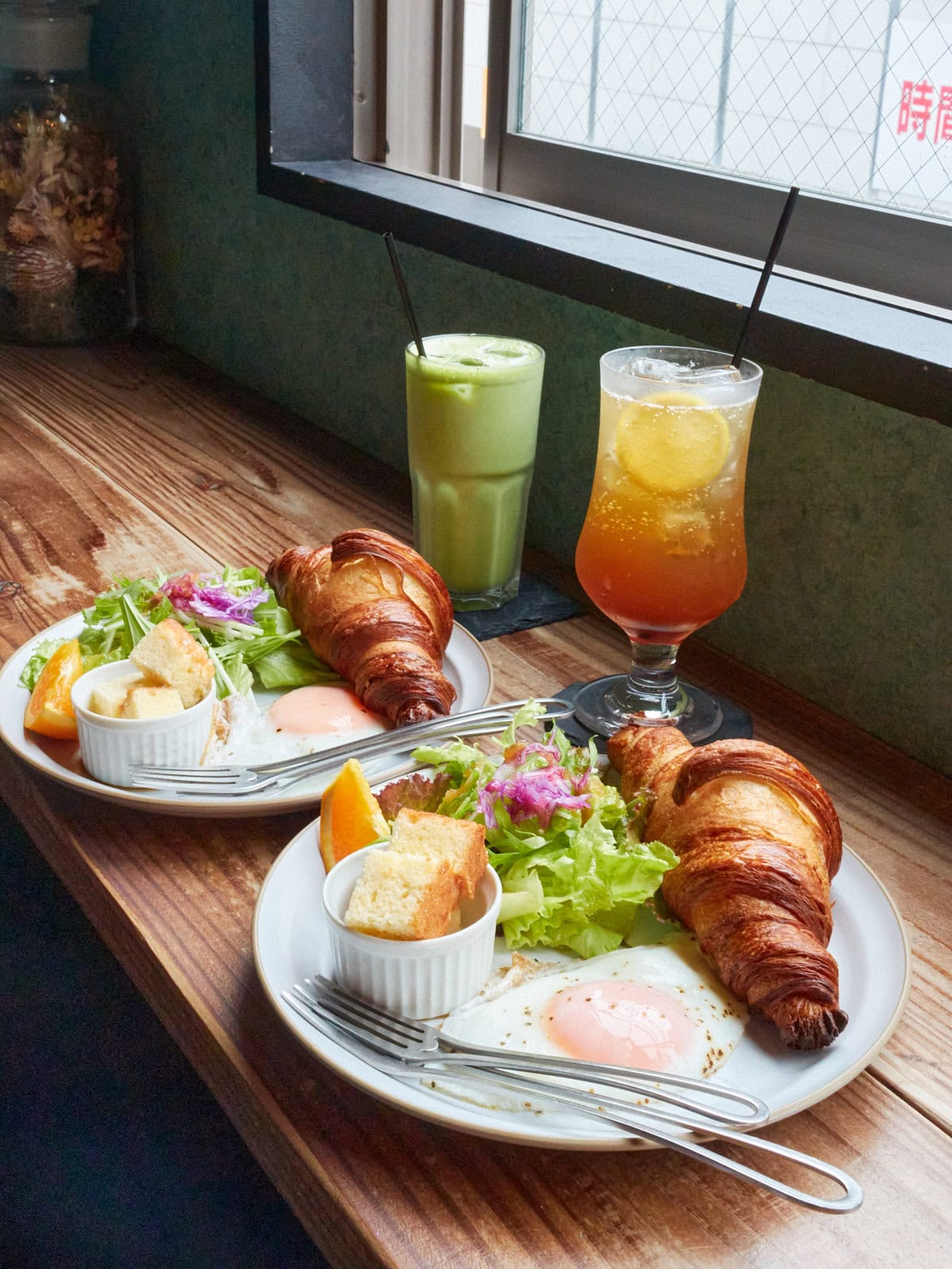 2 morning sets with croissant, a fried egg, salad, orange slice, pound cake cubes, and drinks