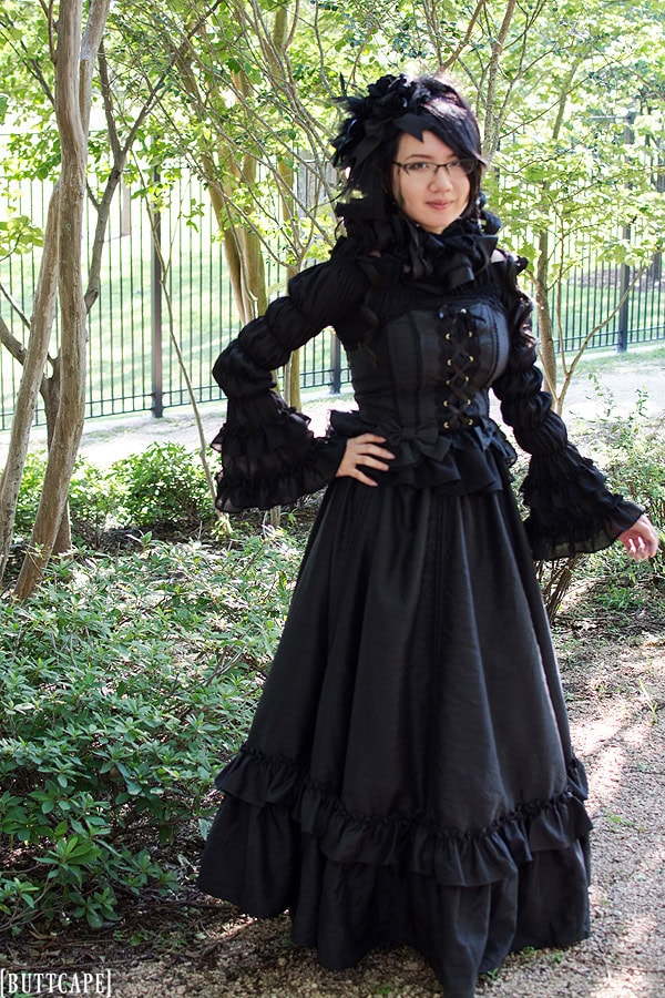 All black gothic girl standing in park with long ruffle sleeves, corset, and long skirt.