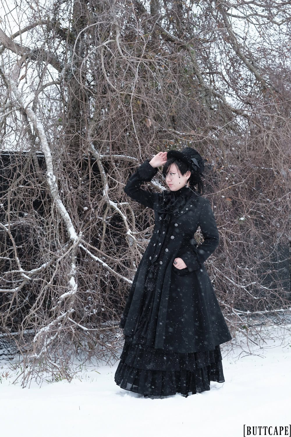 Victorian gothic person wearing all black outfit standing in front of dead tree.