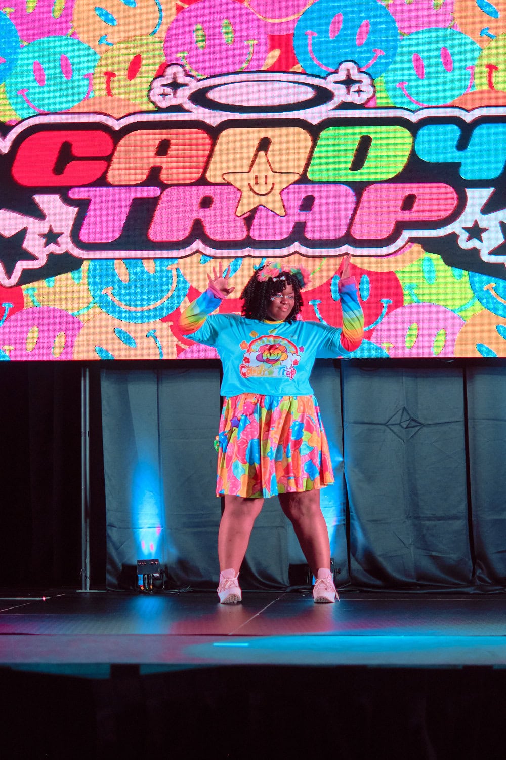Candy Trap model #10 wears logo print t-shirt with striped rainbow sleeves. They also wear a knee length colorful skirt.