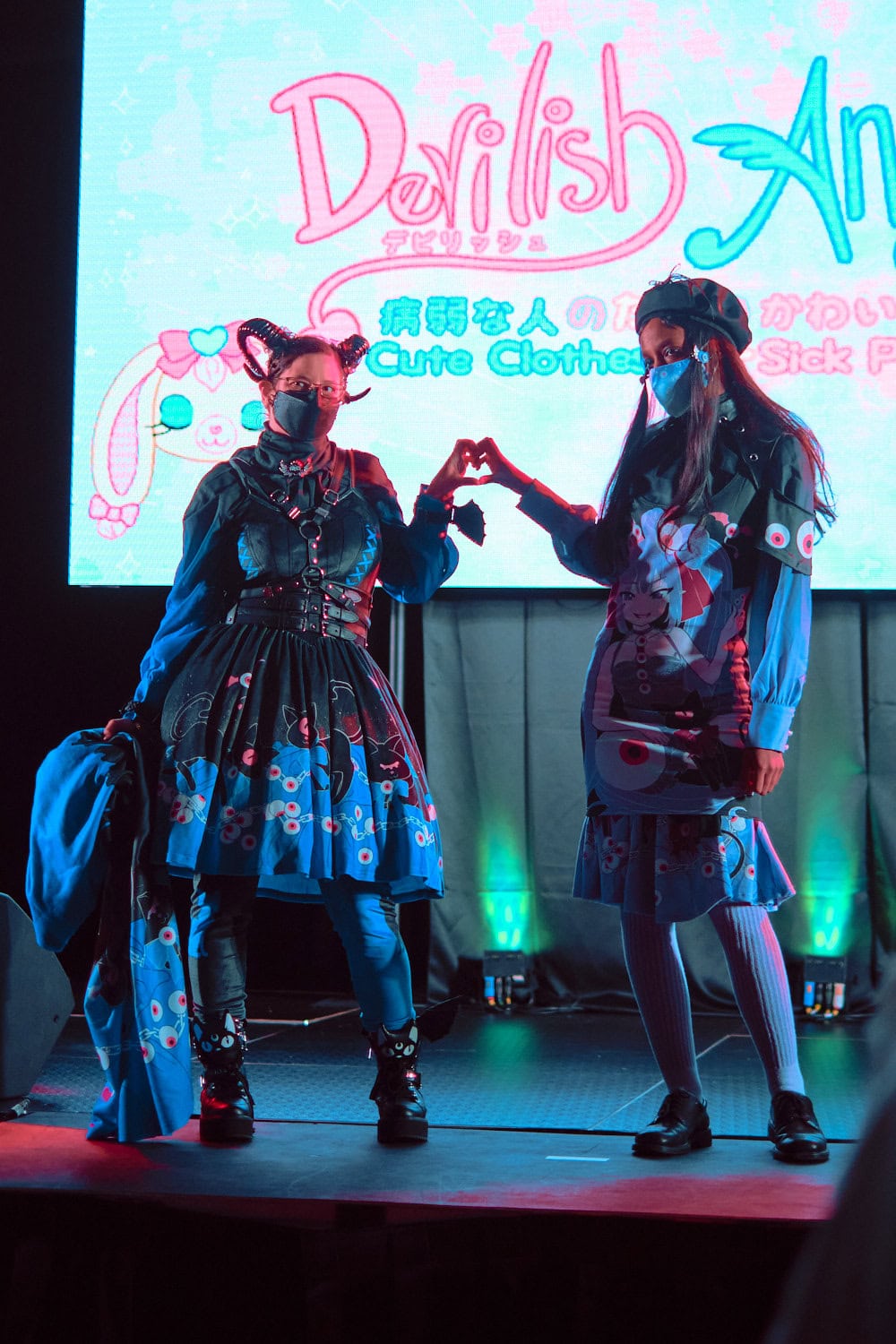 Devilish Angel models #2 and #3 stand next to each other wearing dark gothic eyeball themed motifs. Model on left wears gothic lolita style with devil horns. Model on right wears a casual beret, long sleeve shirt, and knee length pencil skirt.