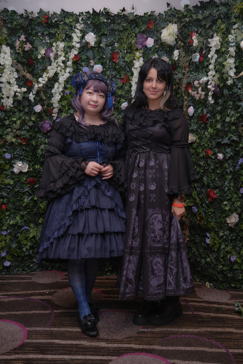 Two gothic lolitas standing in front of floral wall.