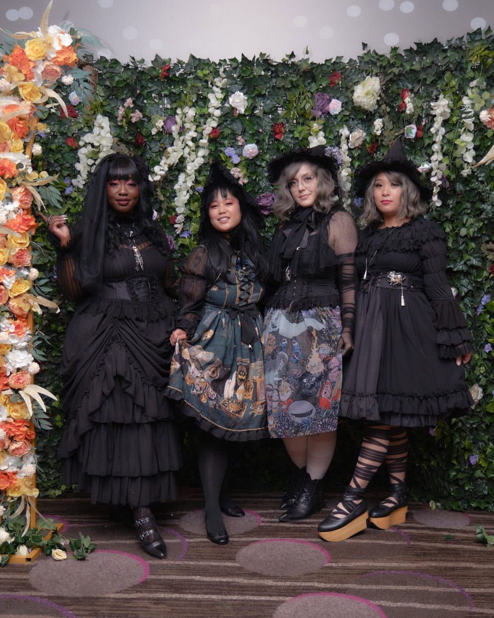 Four gothic lolitas standing in front of floral wall.