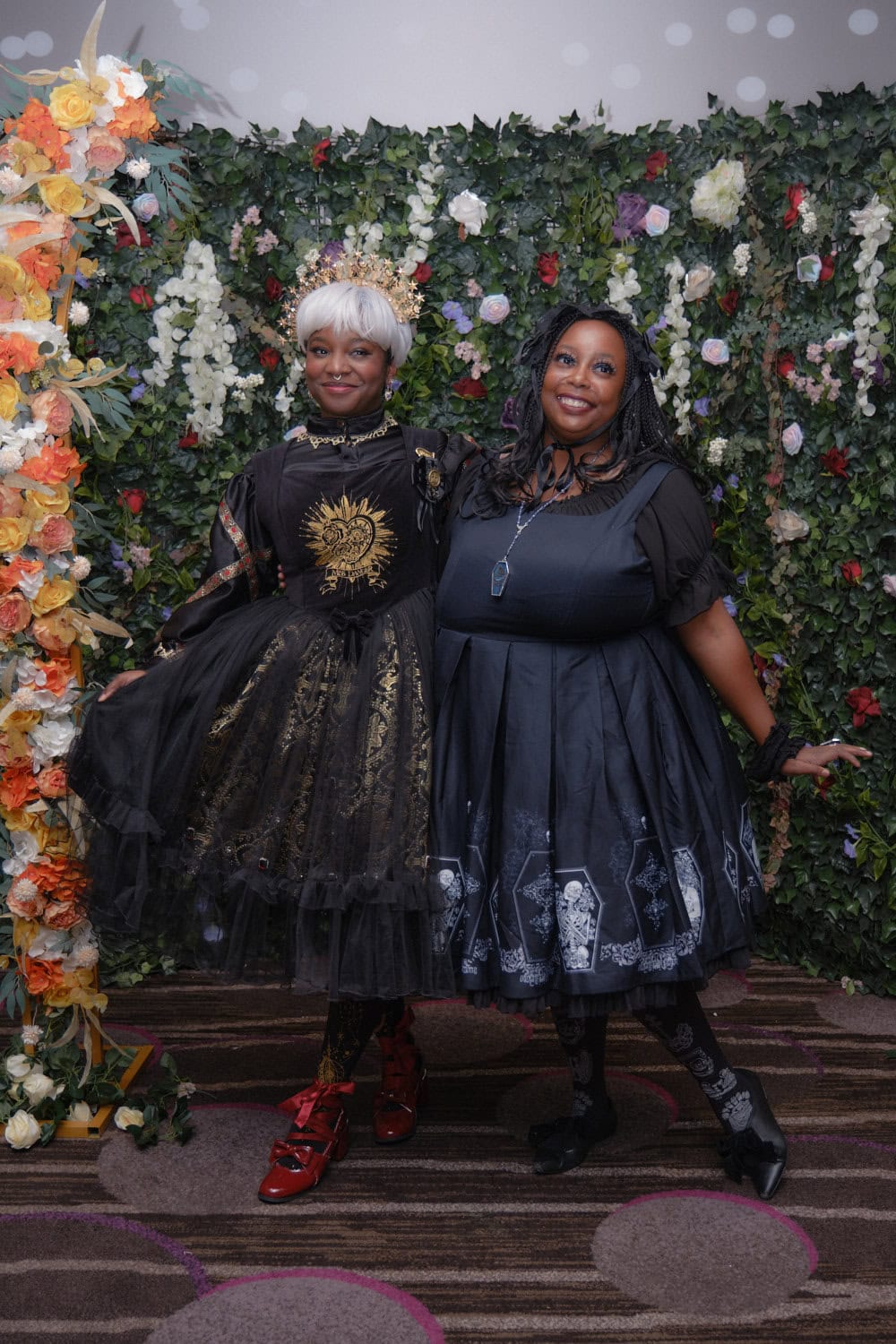 Two gothic lolitas standing in front of floral wall.