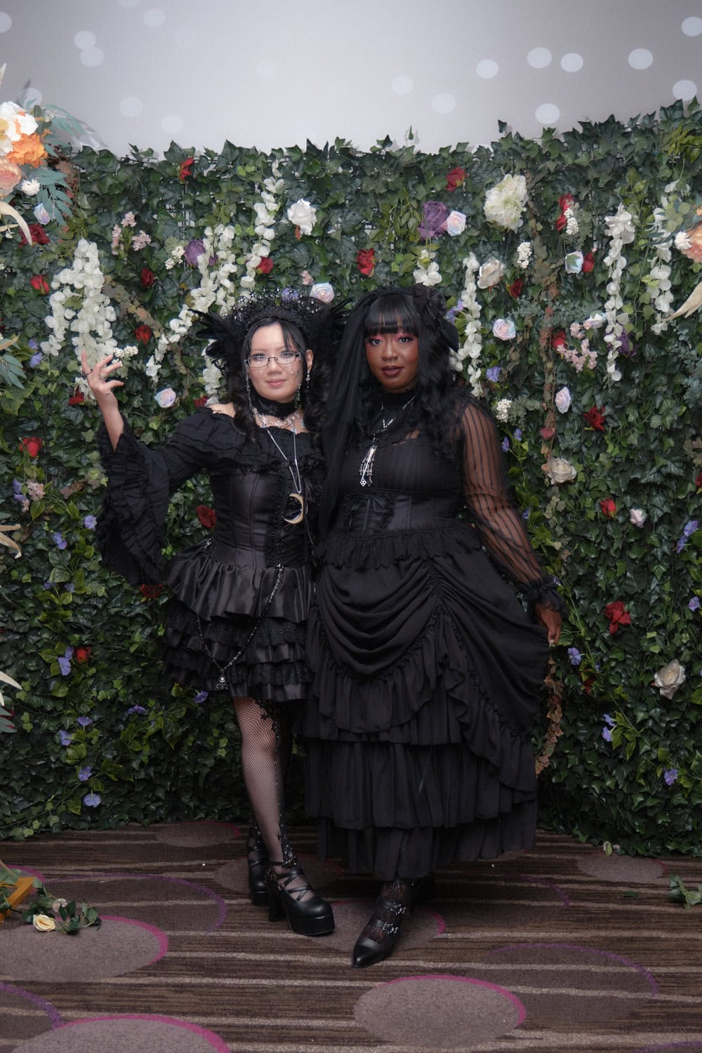 Two gothic lolitas standing in front of floral wall.