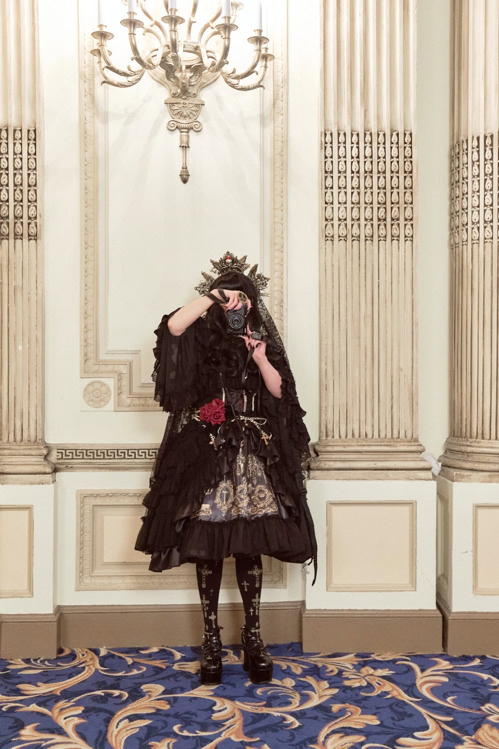 Gothic lolita holding camera in front of face wearing black and gold print dress.