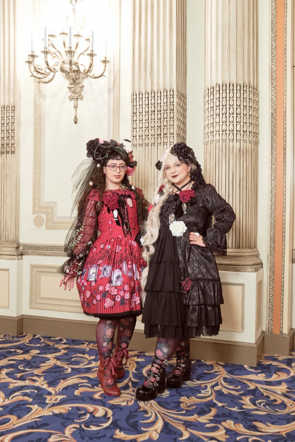 Two gothic lolitas with rose accents standing next to each other. Left is wearing red x black and right is wearing black x red.
