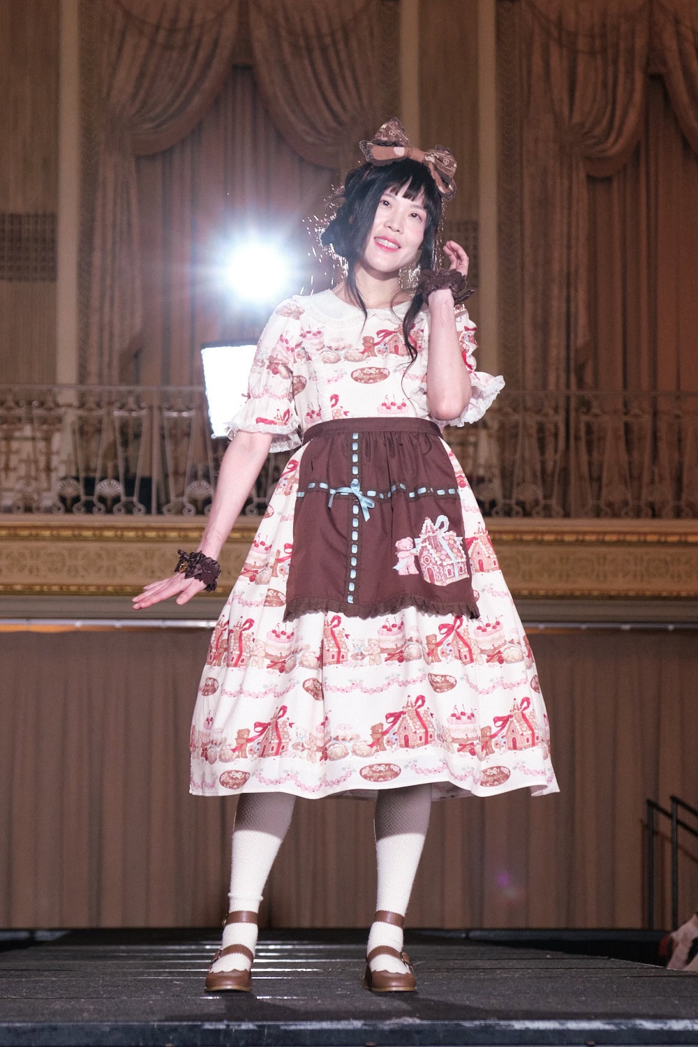 Model 12 wearing pink gingerbread themed sweet lolita short dress with brown apron by Axes Femme Kawaii.