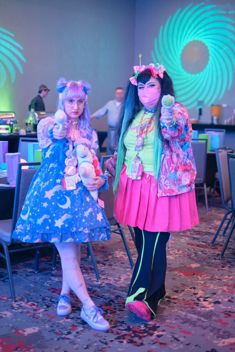 Two friends wearing space themed outfits standing next to each other pointing toy water guns at camera.