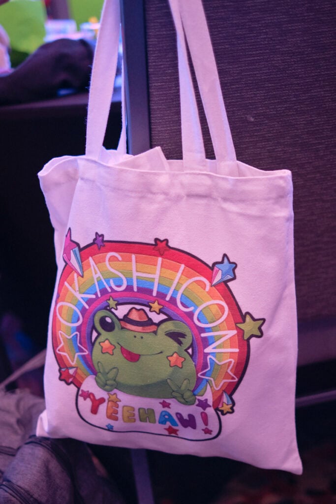 Tote bag featuring a frog sitting under a rainbow. The rainbow has "Okashicon" text on it. The frog is making peace signs with hands with a speech bubble saying "Yeehaw!"