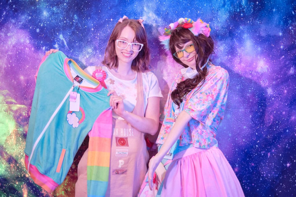 Raffle prize winner holding pastel jacket with striped rainbow sleeves standing next to the designer of Devilish Angel,