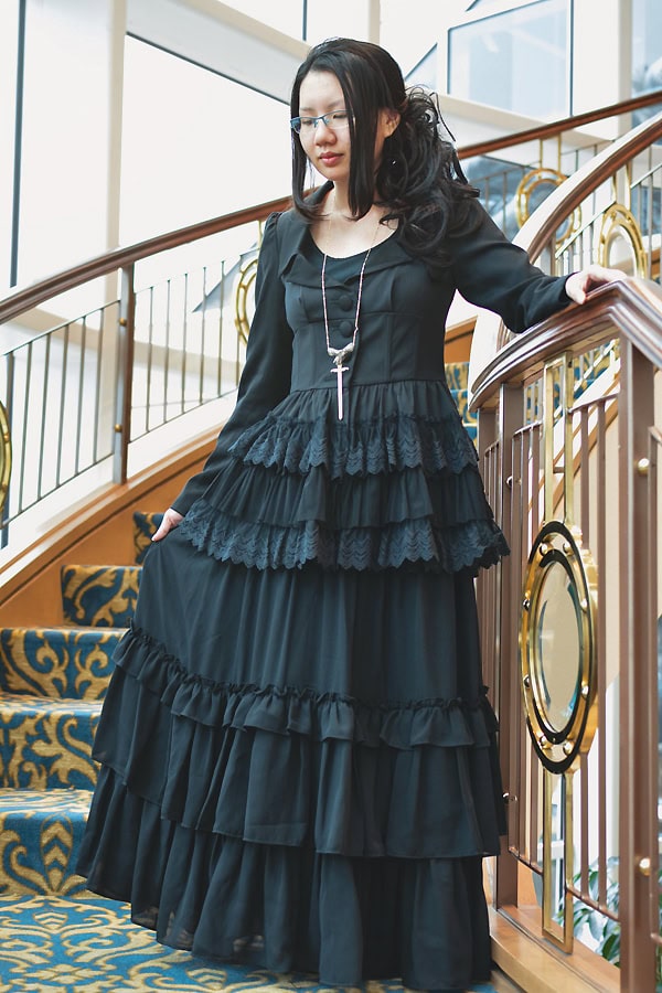 Stephano wearing an all black outfit on a curved staircase. The outfit consists of a long sleeve jacket with lace hem details and a long floor length tiered chiffon skirt.