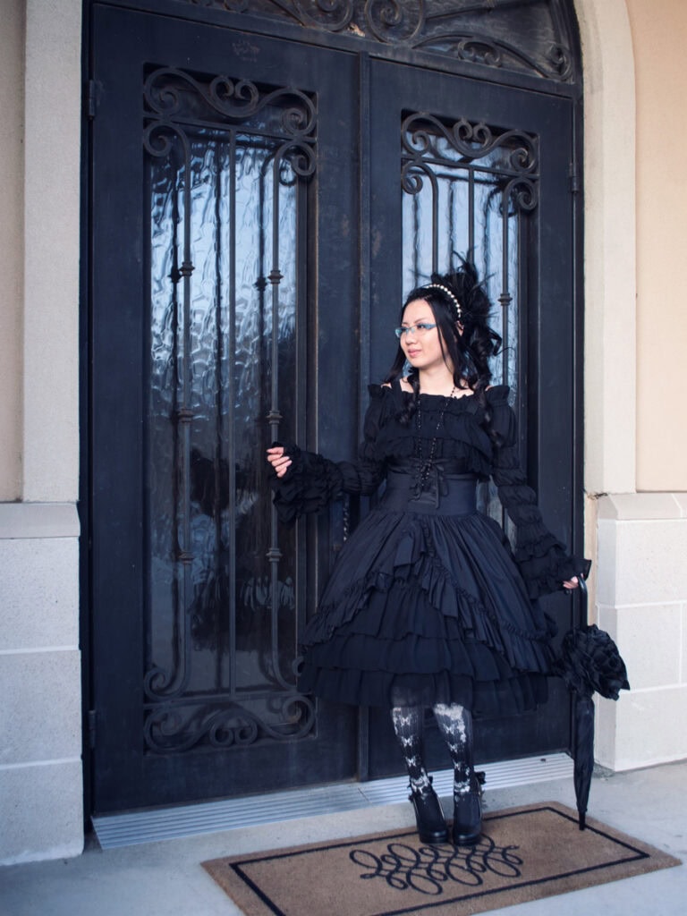 Full body portrait of Stephano wearing gothic lolita in front of an ornate black door. Stephano is facing towards the right and holding a folded parasol like a cane in her left hand. Her right arm is raised at a right angle away from her body,