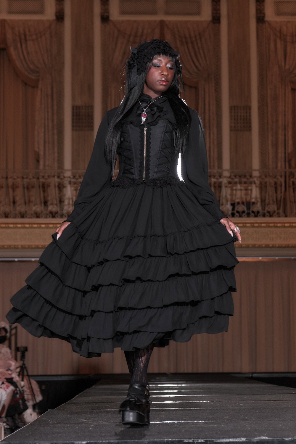 Model 4 wearing an all black aristocratic outfit. Outfit consists of a long sleeve blouse, bustier, and long solid tiered skirt. Model is mid-walk with the skirt flowing in the air.