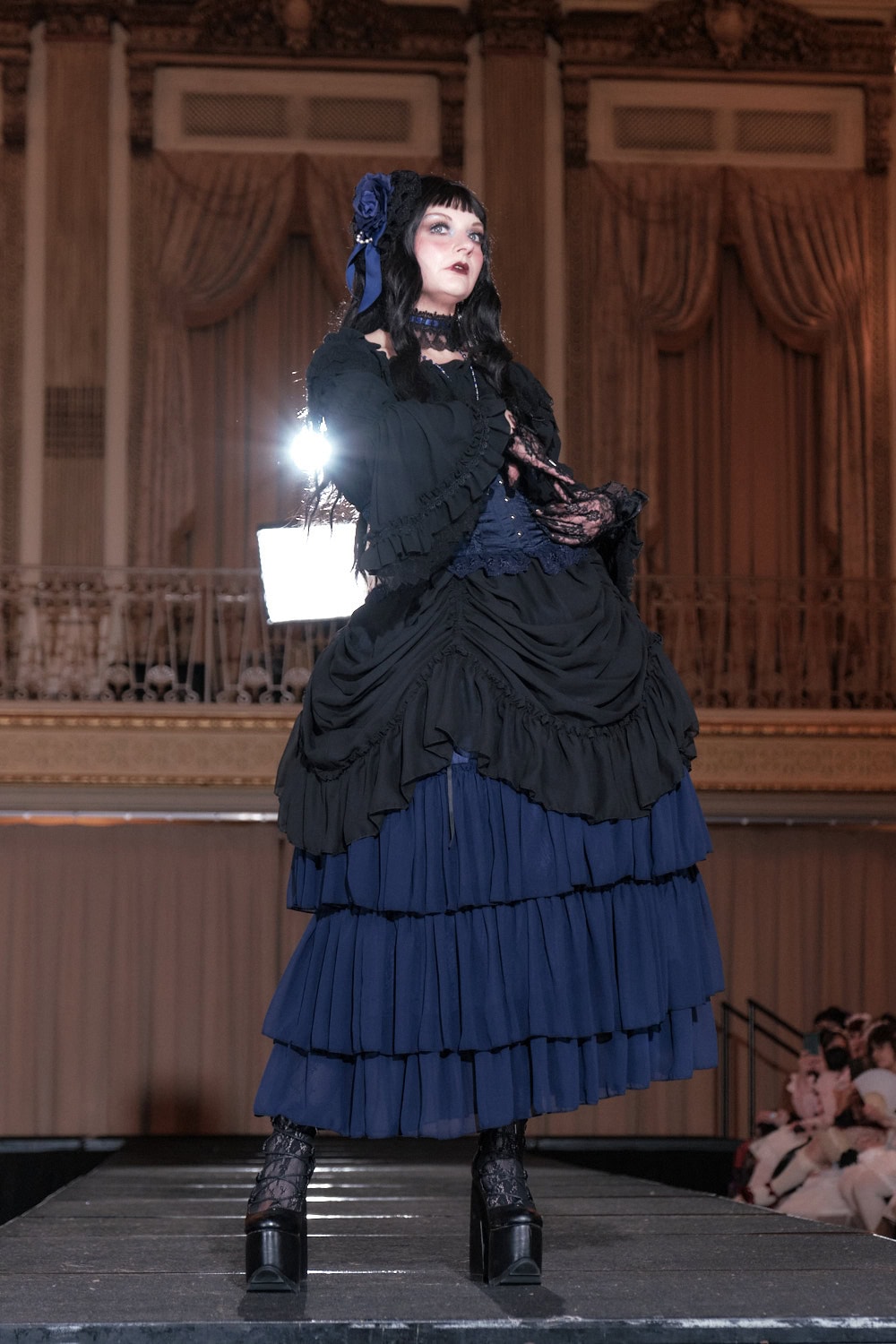 Model 5 wearing a black and navy gothic lolita outfit. The dress is black and has a bustle skirt drawn up to reveal the navy skirt portion. Model is wearing a matching rose headband and choker.