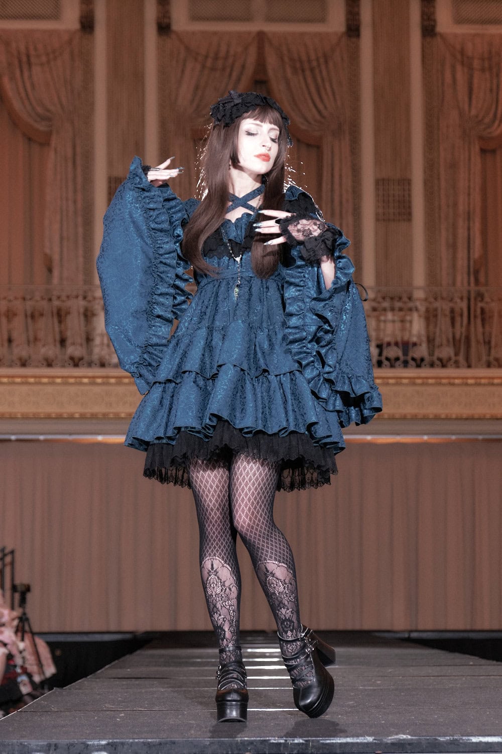 Model 8 wearing turquoise and black jacquard thigh length dress. The model displays the dramatic sleeves draped on her arms.