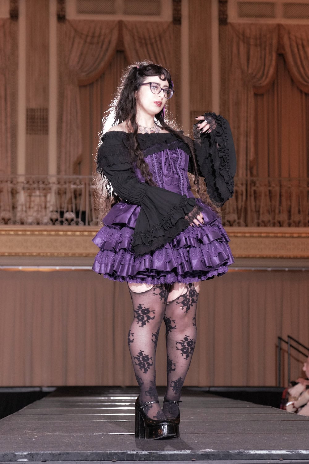 Model 9 wearing black and purple gothic lolita outfit. The thigh length dress is purple paired with an off shoulder black blouse and lace stockings.