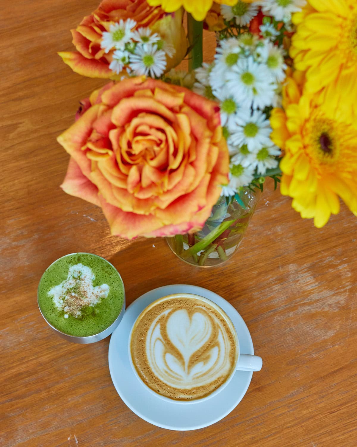 orange roses, white flowers, and yellow flowers framing a matcha cardamom latte and coffee cardamom latte