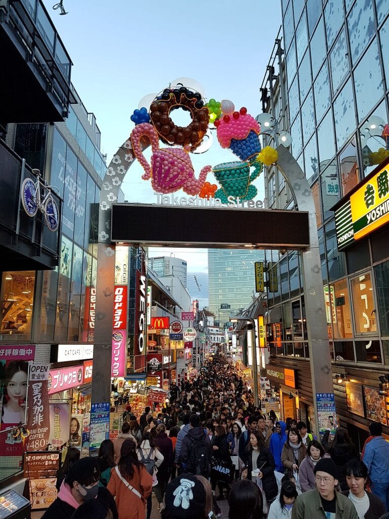 Takeshita Street entry way arch decorated with cute donut and tea cup balloons. Lots of tourists are walking up and down the street.