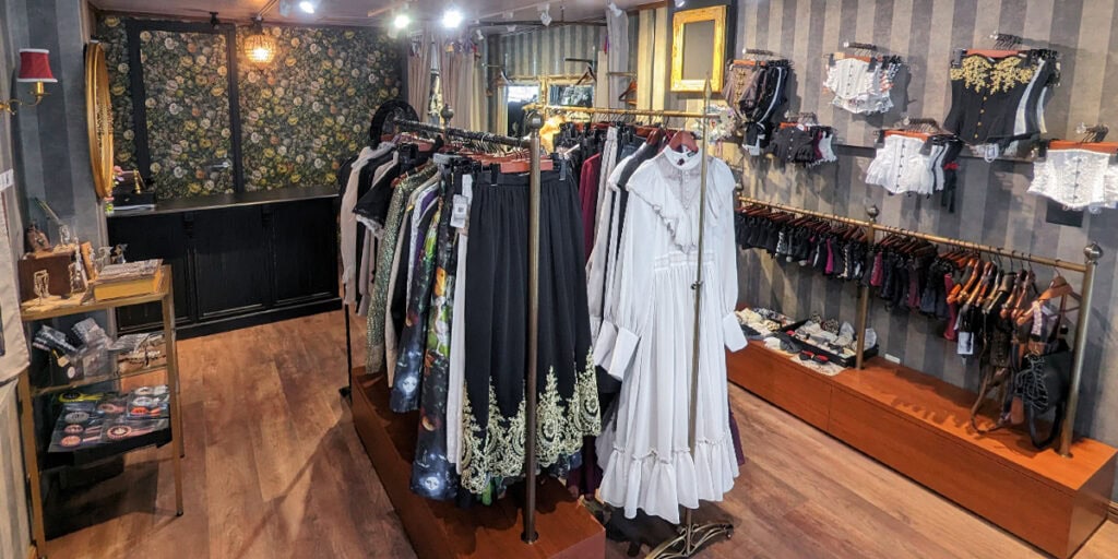 Interior of Abilletage shop. Clothing racks are filled with long dresses and skirts. The walls are lined with corsets of many designs.