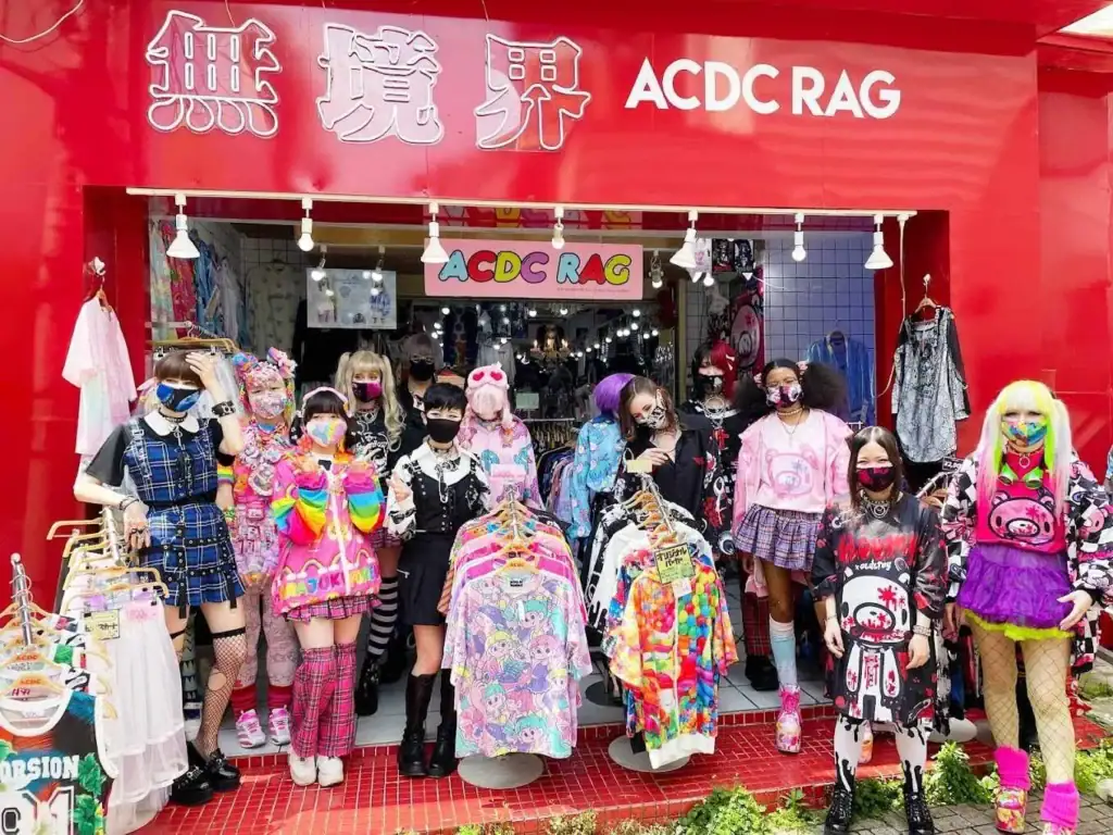 ACDC Rag Harajuku shop front. The building has bold reed walls. Graphic T-shirts are hanging inside and outside the shop. About 10 people dressed in kawaii fashion stand in front of the shop.
