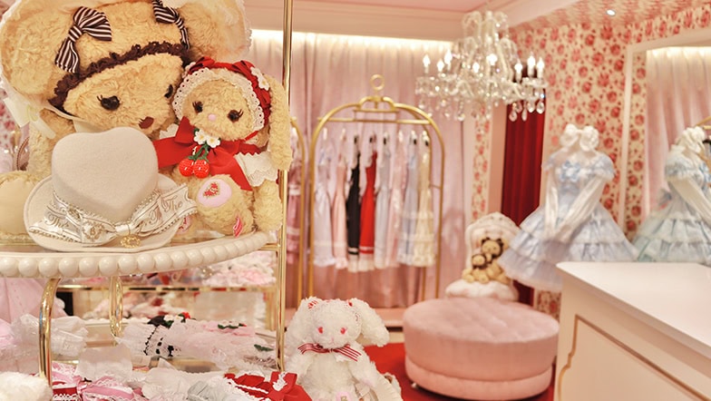 Shop interior of Baby, the Stars Shine Bright's Harajuku store. The foreground features 3 bags of their mascots usakumya and kumakumya. The background has lolita dresses and a chandelier.