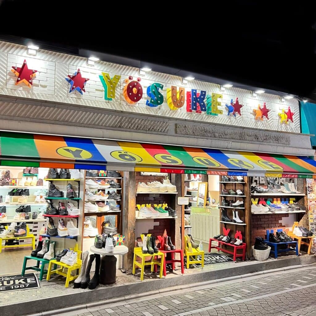 Front of the Yosuke shoes shop on Takeshita Street. The photo is taken at night and the shop is lit up by lights. Many different styles of platform shoes are displayed inside and outside of the store.