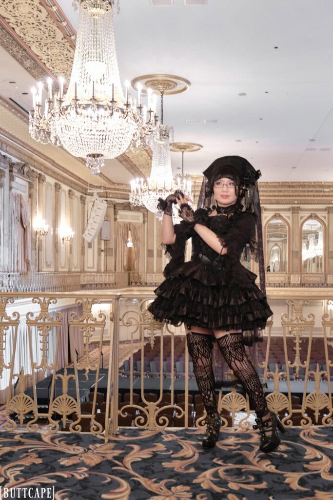 Full body photo Stephano wearing an all black gothic lolita outfit for day 1 of The Great Lace featuring pieces from Atelier Pierrot mostly. She is standing in front of several chandeliers.