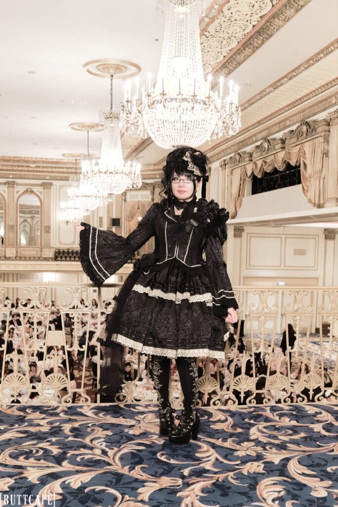 Full body photo of Stephano's outfit for the Great Lace day 2. The outfit is a black and gold hime lolita outfit with rose accents. She ins standing in front of a row of chandeliers.