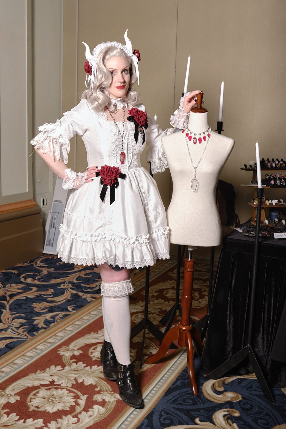 Neant Glass designer Kei wearing all white lolita outfit with red rose accents. She is posing with a mannequin wearing two of her necklaces.