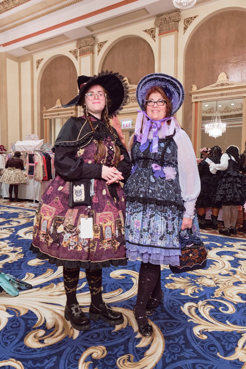 Two attendees standing next to each other. The left attendee wears a magic inspired lolita outfit with a witch hat, capelet, and magic themed print by Metamorphose. The right attendee wore a black and purple hydrangea themed classic lolita print by Haenuli.